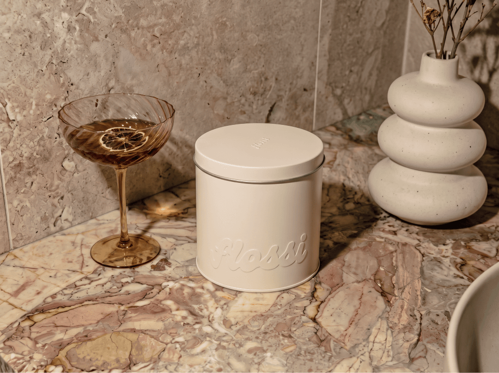 The cream Flossi tin, wordmark embossed on the front, on a marble bathroom counter